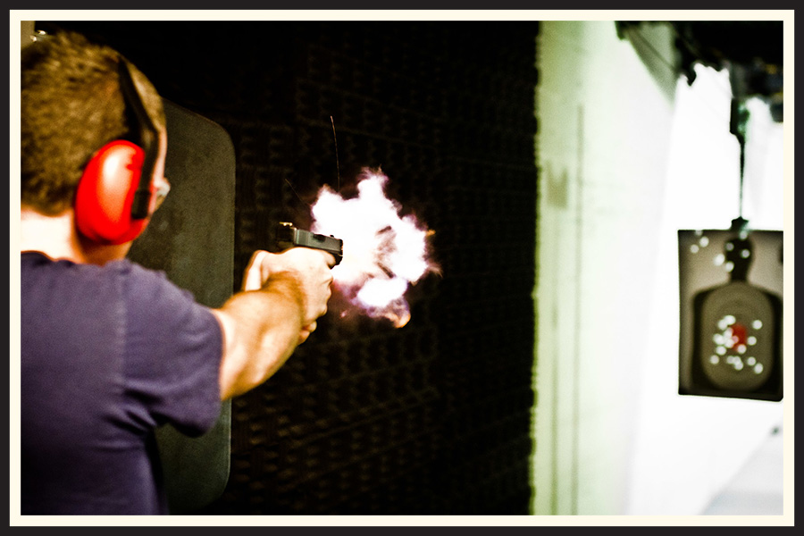 A man fires a gun in a shooting range, the explosion caught by the slow shutter speed of the photo.