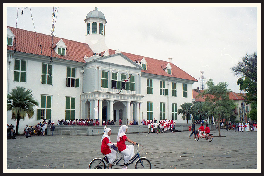 Two people in red and white ride a tandem bicycle, taken on Kodak Colorplus 200 color film.